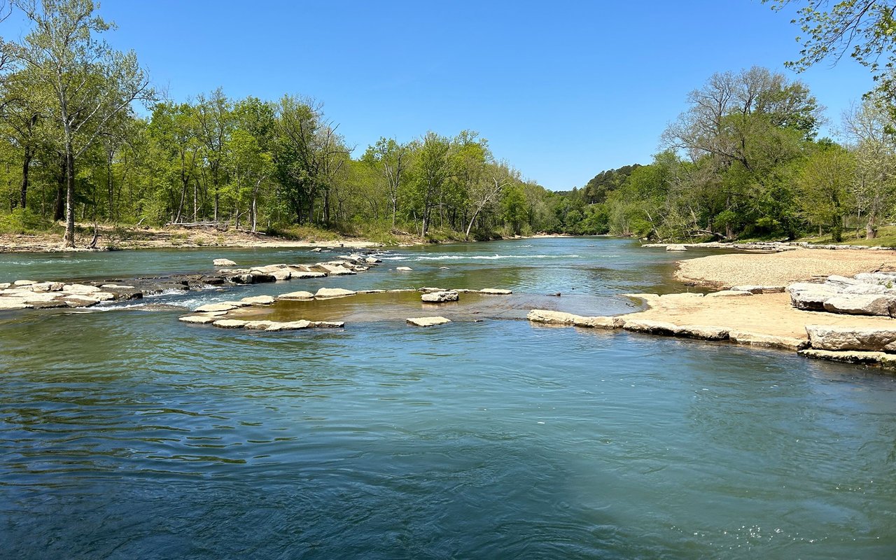River flowing over rock shelves between wooded banks and a sandy shore.