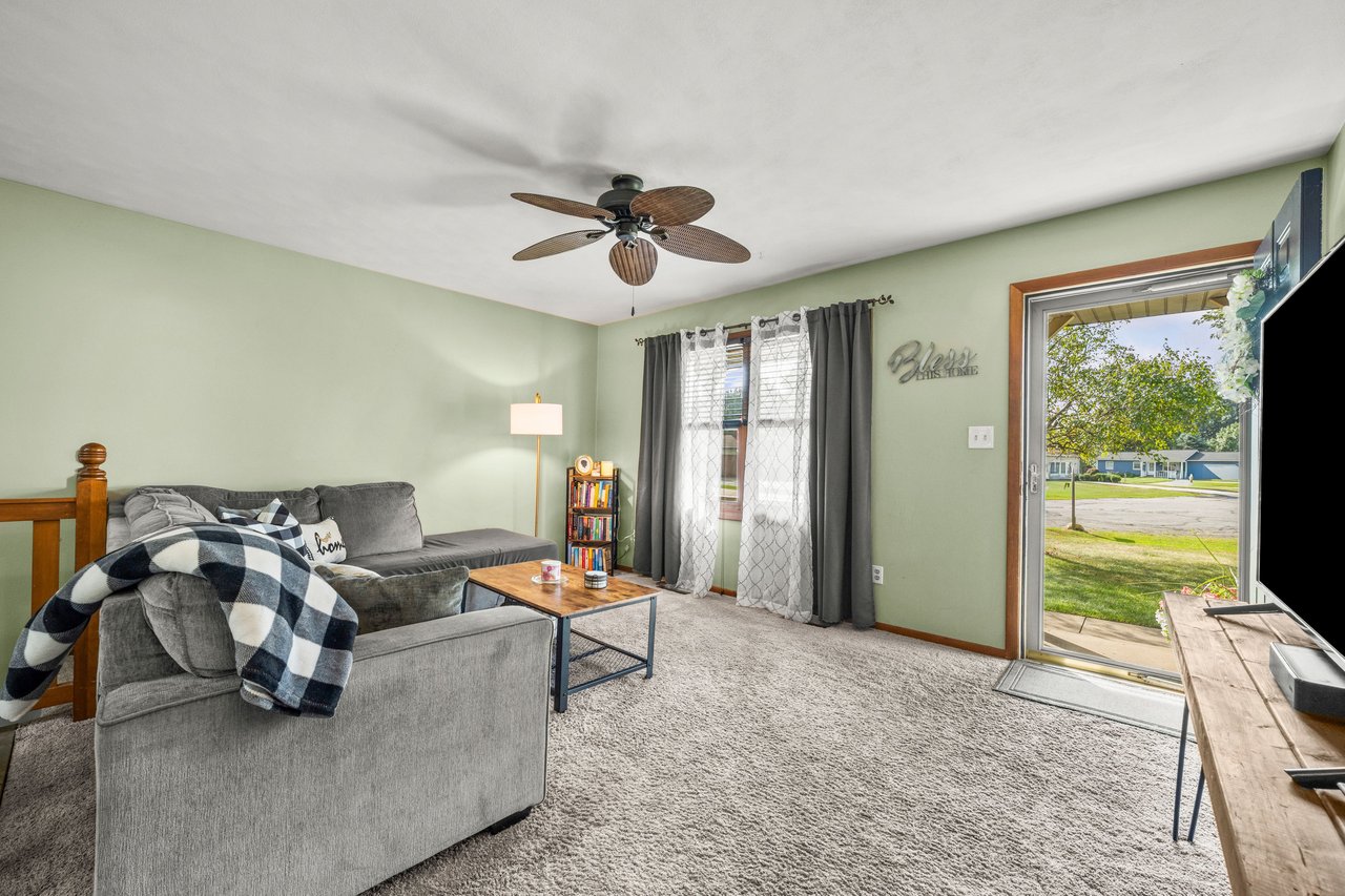 Furnished living room with a gray sectional, ceiling fan, and open patio door overlooking a lawn.