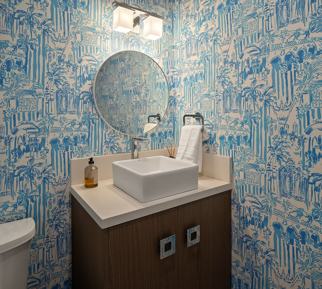 Bathroom vanity with a vessel sink, round mirror, and blue illustrated wallpaper.