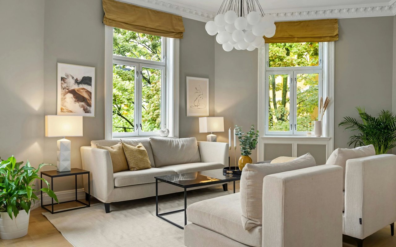 Furnished living room with cream seating, tall leafy-window views, and a white globe pendant light.