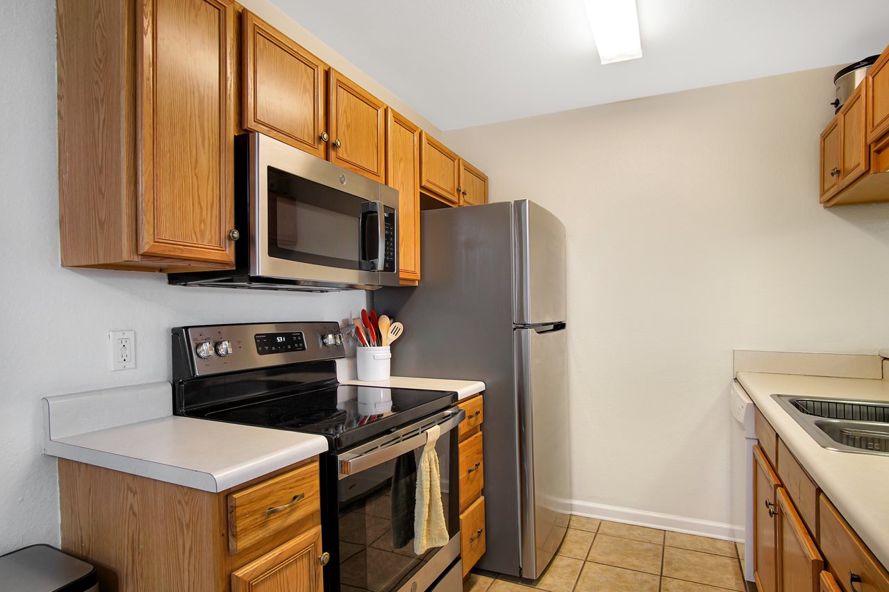 Kitchen with wooden cabinets, stainless steel appliances including a stove, microwave, and refrigerator. Countertops hold a utensil jar, creating a warm, functional space.