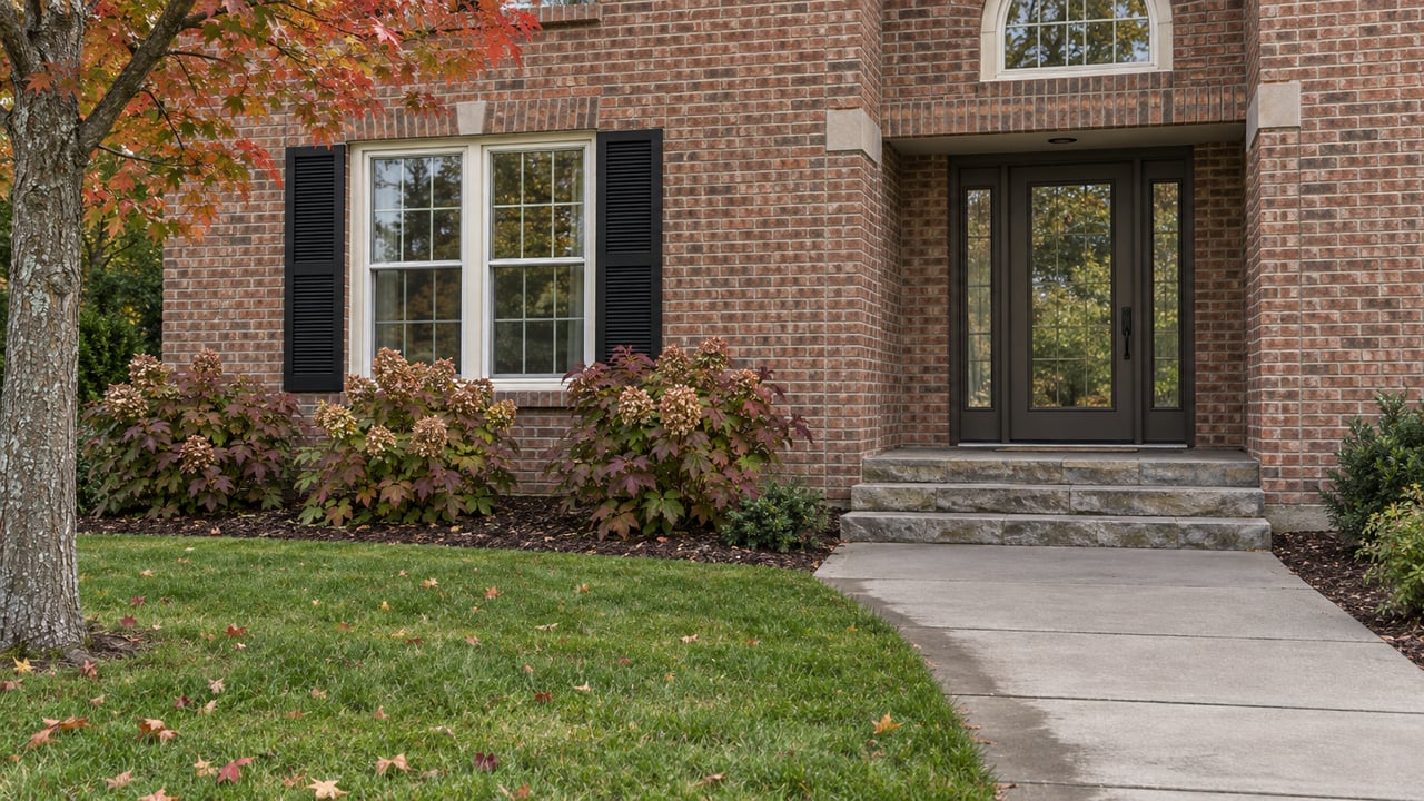 Brick home with a bronze glazed entry, black shutters, oakleaf hydrangeas and a broad lawn beside the front walk.