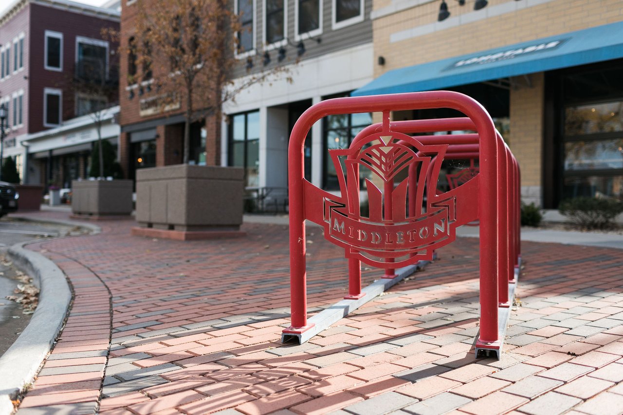 A red metal bike rack featuring the Middleton logo on a brick sidewalk in front of downtown storefronts in Middleton, Wisconsin.