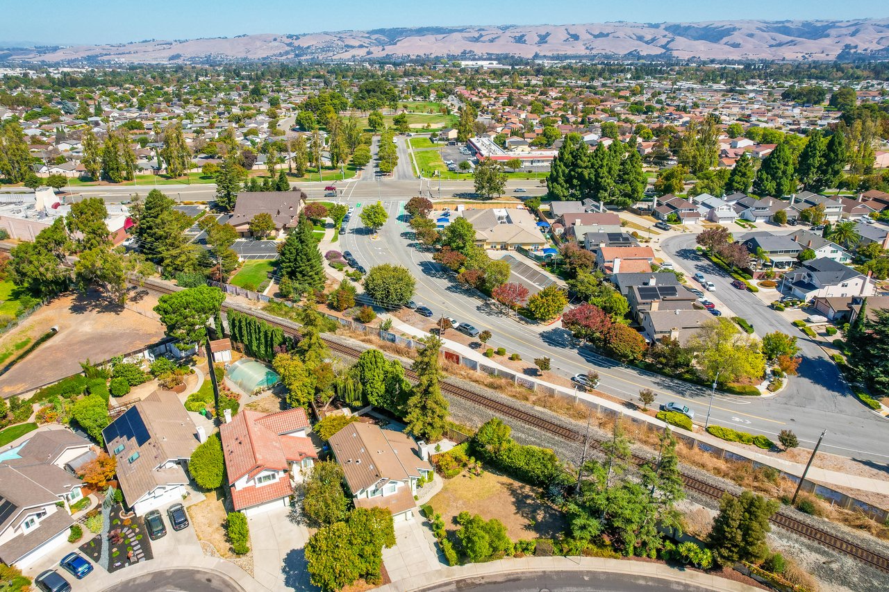 Aerial view of a residential neighborhood with a rail line, green park, and distant hills.