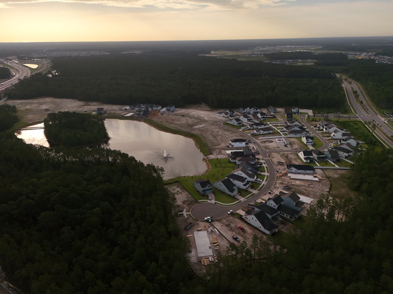 Aerial view of homes under construction around a pond, with a fountain and surrounding woodland.