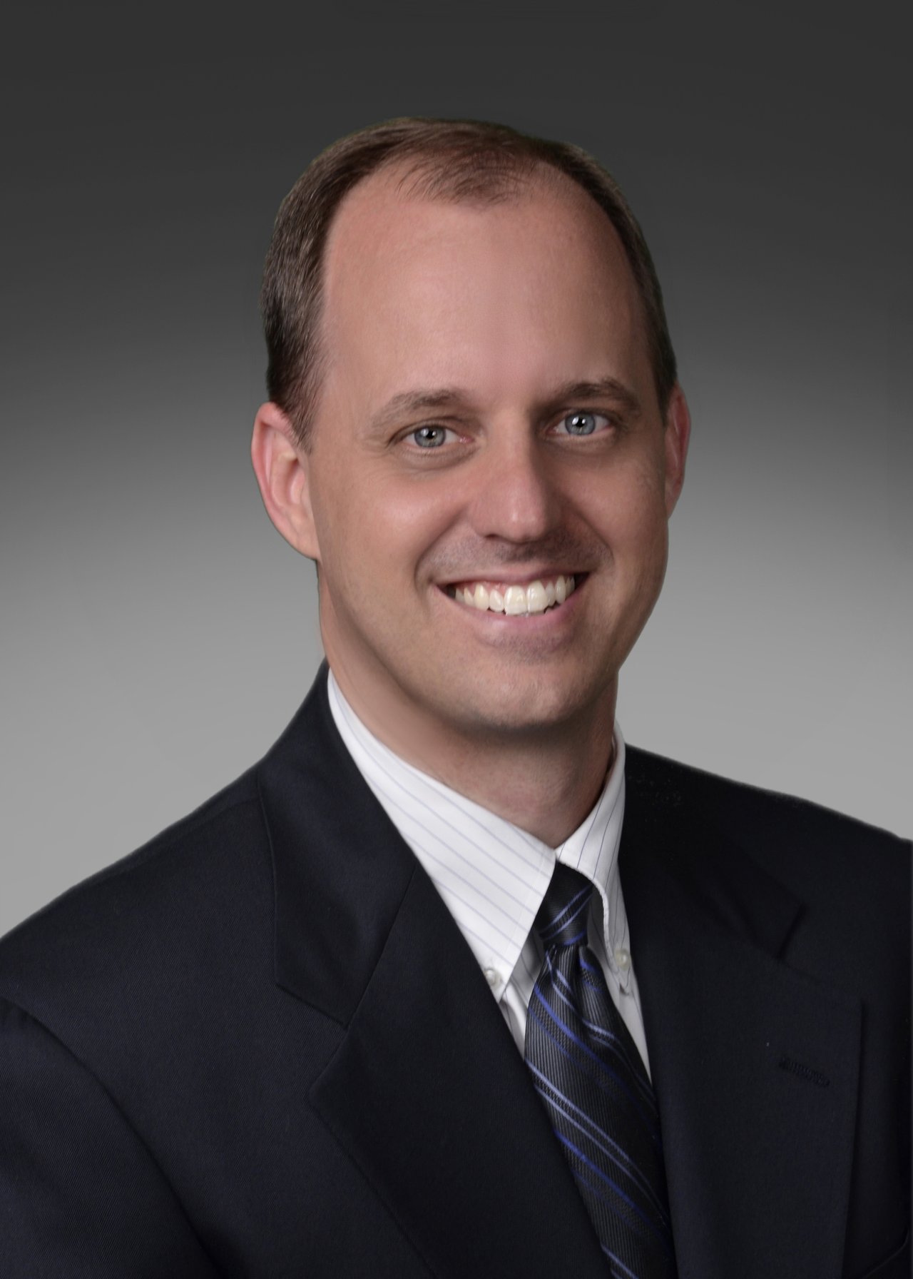 Smiling man in a dark suit and striped tie against a gray studio backdrop.