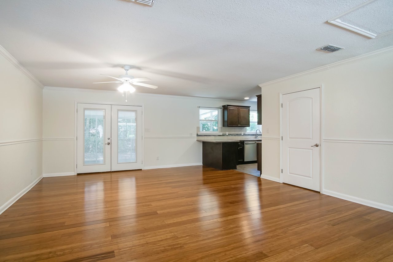 Spacious empty living room with wooden floors and ceiling fan. French doors provide outdoor view. Attached kitchen features dark cabinets and modern appliances, creating a cozy atmosphere.