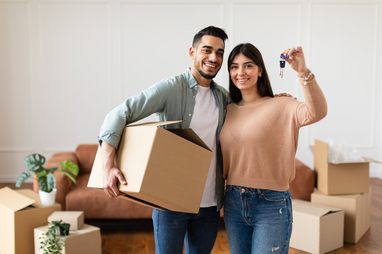 Happy couple moving into their new home in Houston. The woman holds keys while the man carries a moving box, surrounded by unpacked boxes and houseplants—symbolizing the decision between renting and owning a home.
