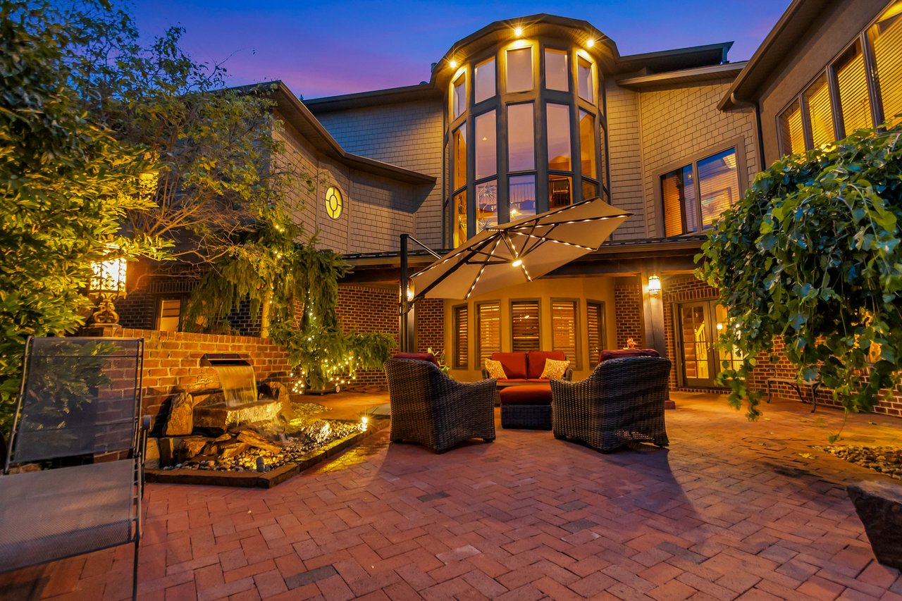 Luxury home courtyard patio with wicker seating, a water feature, and illuminated bay windows at dusk.