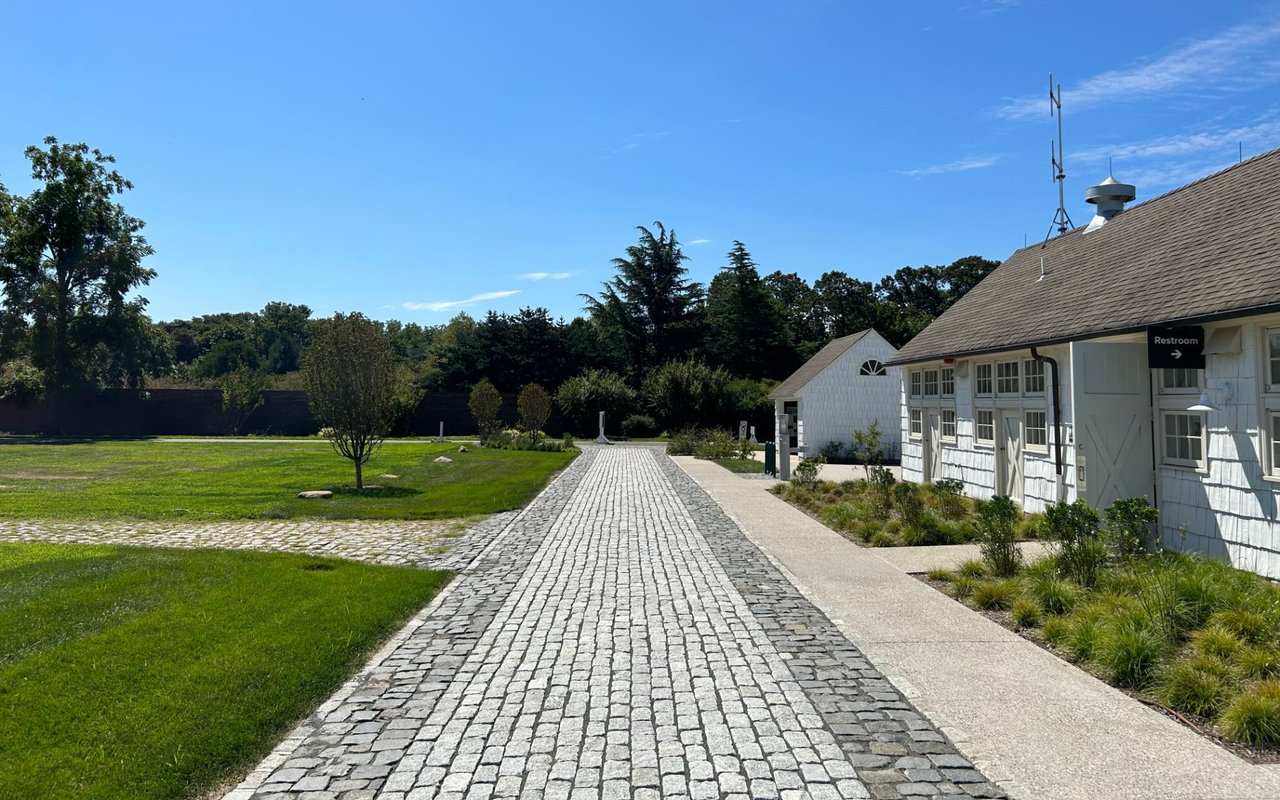 Cobblestone walkway crosses landscaped grounds beside white shingle buildings.
