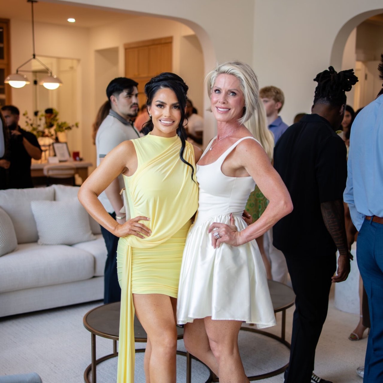 Two women pose at an indoor event, wearing pale yellow and white dresses.