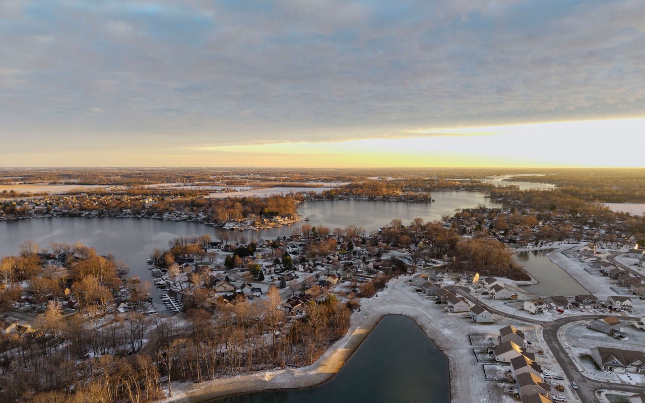 Aerial view of a snowy lakeside neighborhood at sunset, with homes lining the shore.