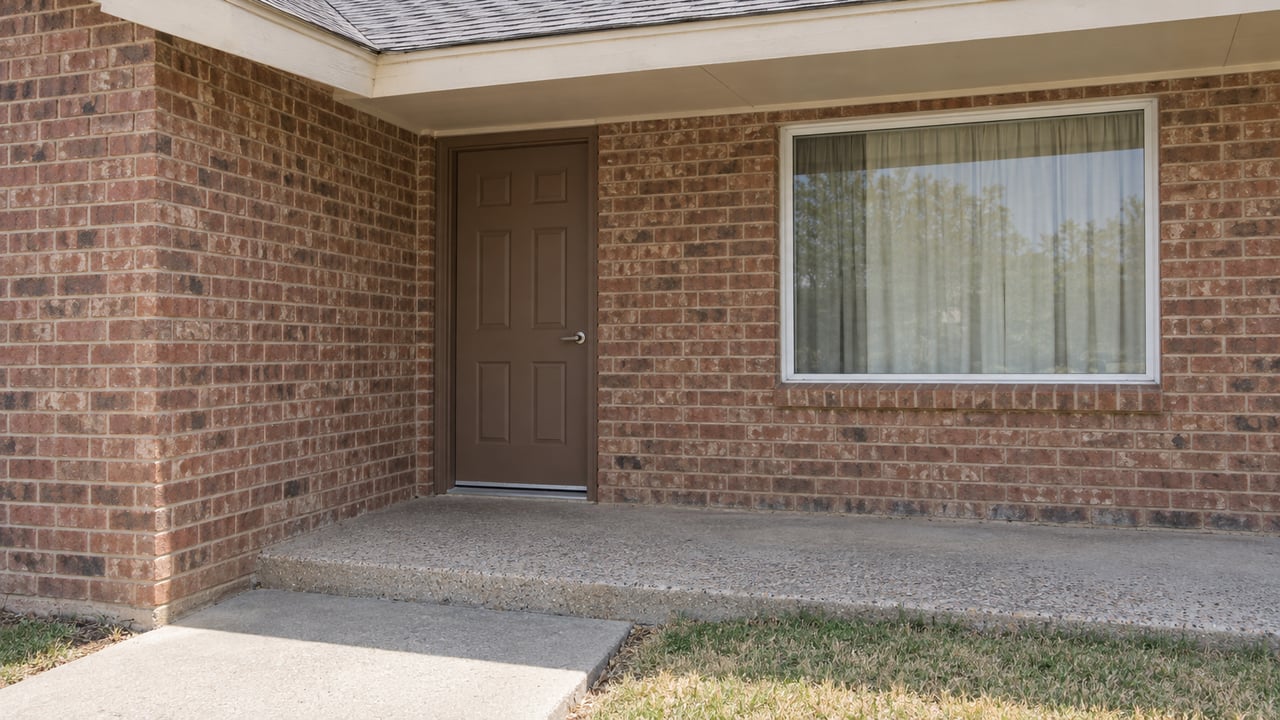 Red-brown brick entry with a closed brown door, cream trim, a picture window and a weathered concrete landing beside short grass.