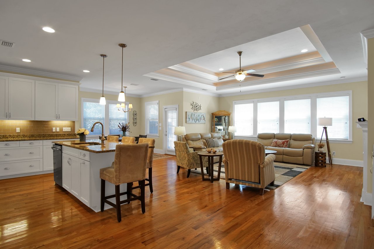 Spacious open-plan living area with wood floors, featuring a modern kitchen with an island, pendant lights, and a cozy seating area with beige sofas.