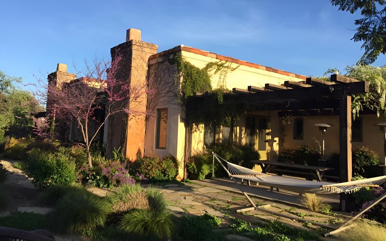 Stucco home with a timber pergola and hammock on a landscaped stone patio.