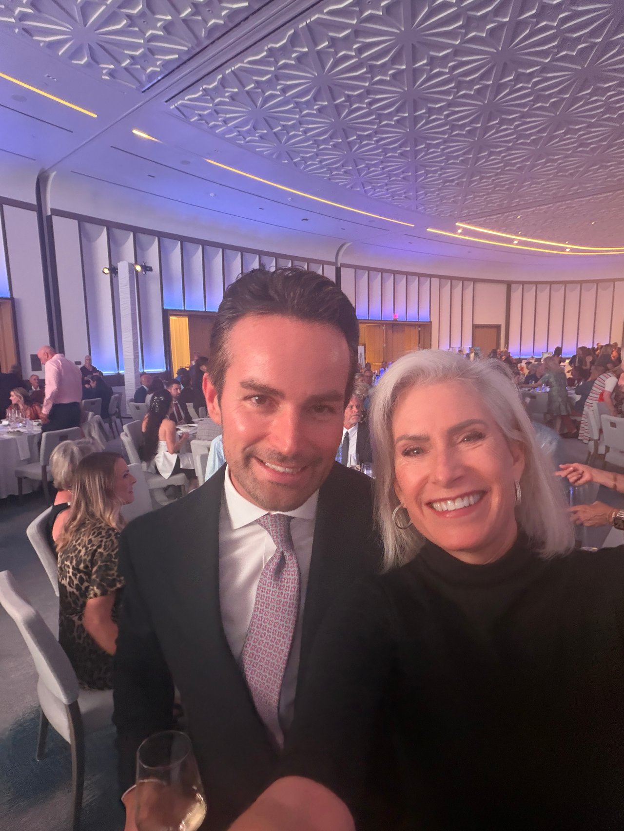 A man in a dark suit and a silver-haired woman take a selfie at a crowded banquet.