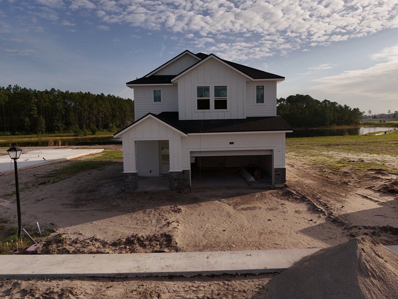Two-story white house under construction beside a pond, with an open garage and unfinished yard.