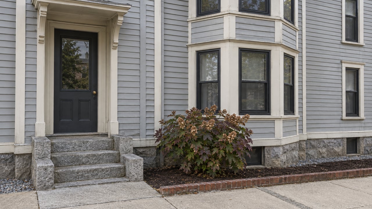 Gray clapboard multifamily facade with stacked bay windows, a sheltered charcoal entry, worn granite steps, and an oakleaf hydrangea in an edged planting bed.