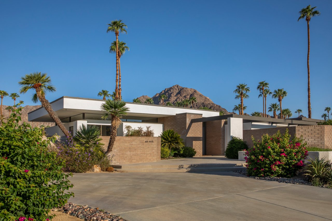 Modernist home with a flat roof, desert landscaping, palms, and a mountain behind it.