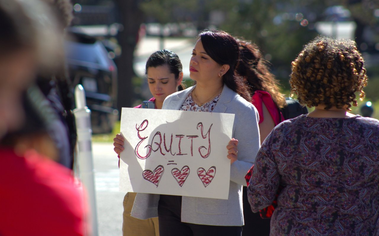 Woman holding an “Equity” sign with three hearts at an outdoor gathering.