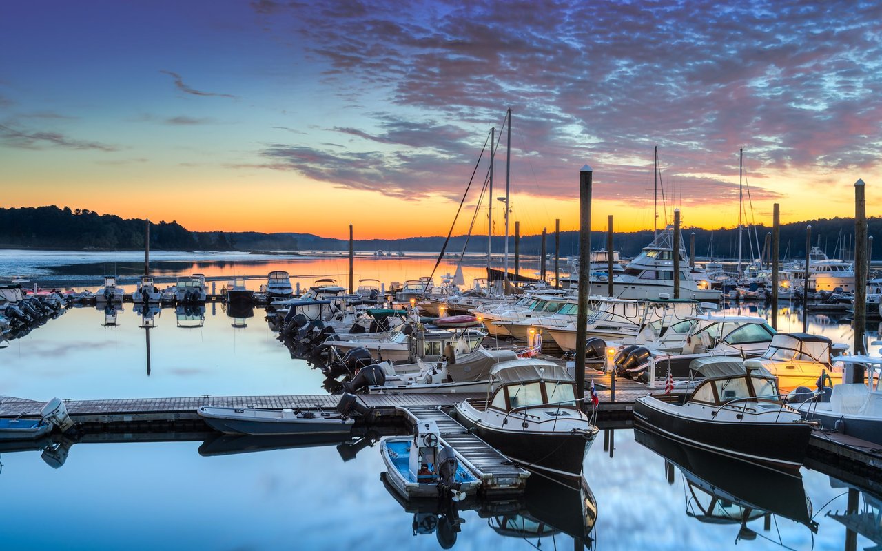 Boats moored at a marina reflect on calm water beneath a colorful sunset sky.
