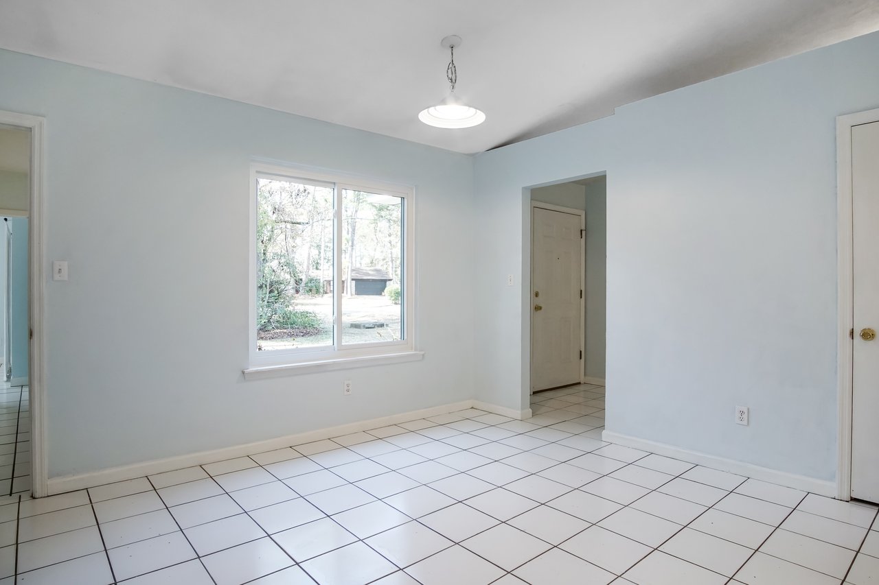 Empty room with pale blue walls and white tiled floor. A large window shows greenery outside. A ceiling light hangs centrally, creating a calm, clean atmosphere.