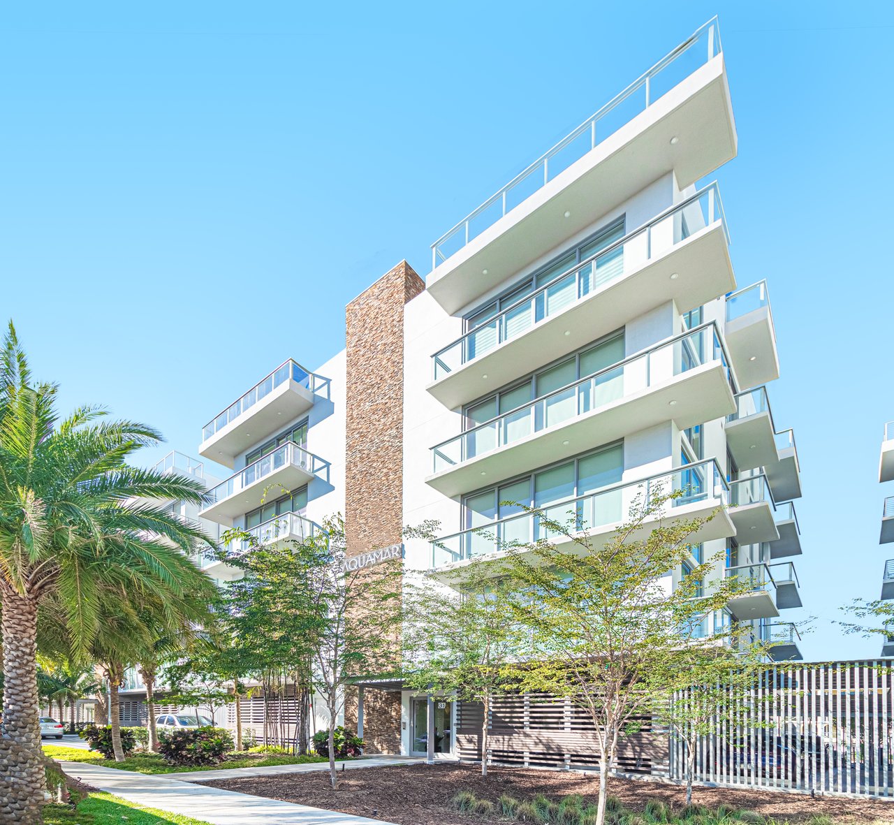 Modern condominium building with glass-railed balconies, a brick accent tower, and palm-lined grounds.