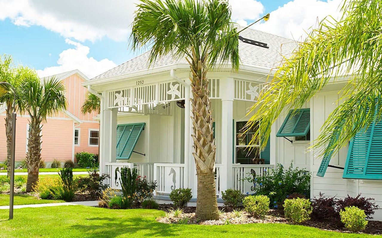 White cottage with teal shutters, a covered porch, and palm trees in the landscaped front yard.