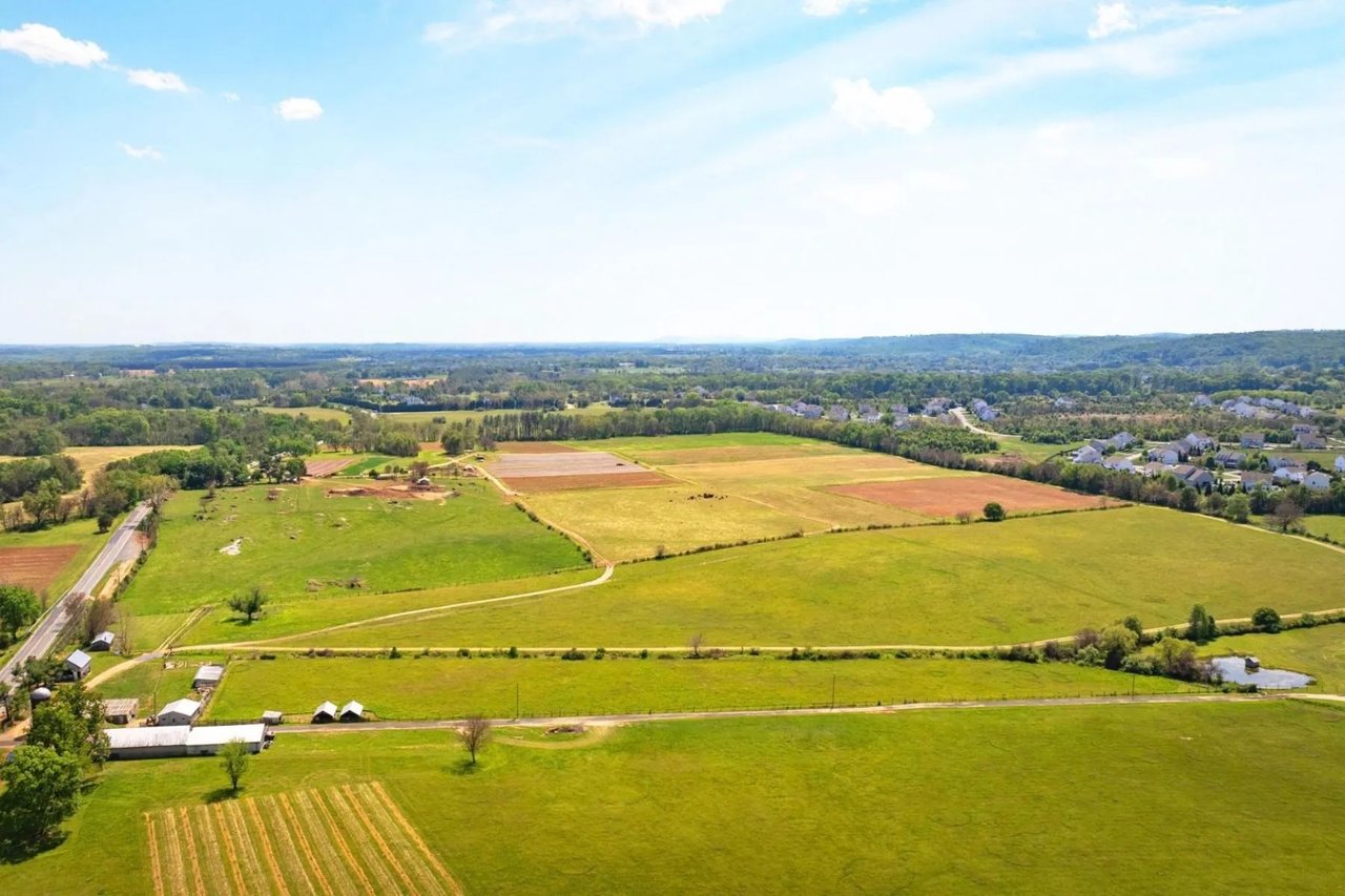Aerial view of green fields and cultivated plots bordered by a road, with farm buildings and distant homes.