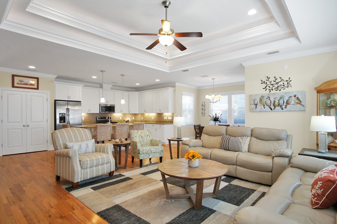 Spacious living room with beige walls, leather sofas, striped and floral armchairs. Wooden furniture, ceiling fan, kitchen in background; cozy and inviting.