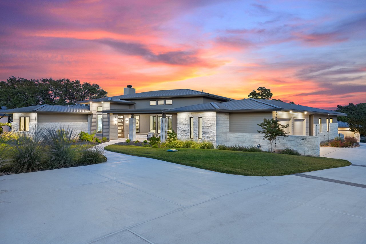 Twilight front elevation of a Hill Country modern home on Caudill Lane in the Bee Caves area of Austin, Texas, framed by mature oak trees along the driveway.
