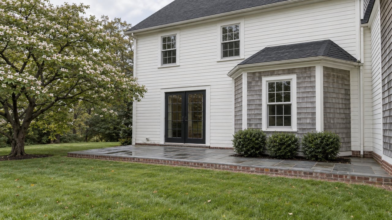 Two-story white clapboard house with a shingled bay, French doors, and a brick-edged stone terrace beside a lawn.