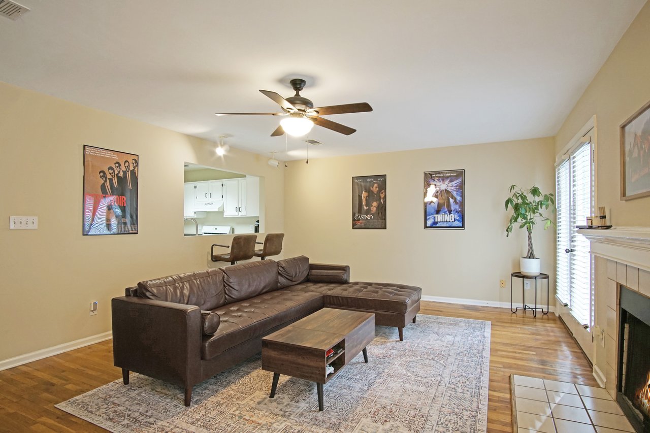 Living room at 529 Collinsford Road, Tallahassee, Florida, featuring a brown leather sectional, patterned area rug, and a modern coffee table. The room includes a ceiling fan with light fixture, fireplace with tile surround, and French doors letting in natural light. A pass-through window opens to the kitchen with bar seating, creating an open-concept feel.