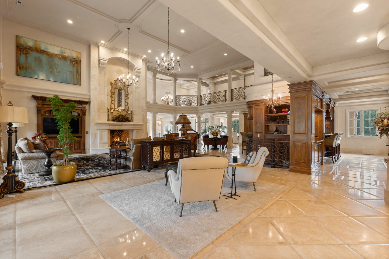 Estate living room with a double-height ceiling, ornate balcony, marble fireplace, and built-in wooden bar.