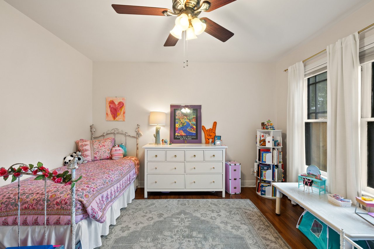 Bedroom with a pink floral bed, white dresser, and large curtained windows.