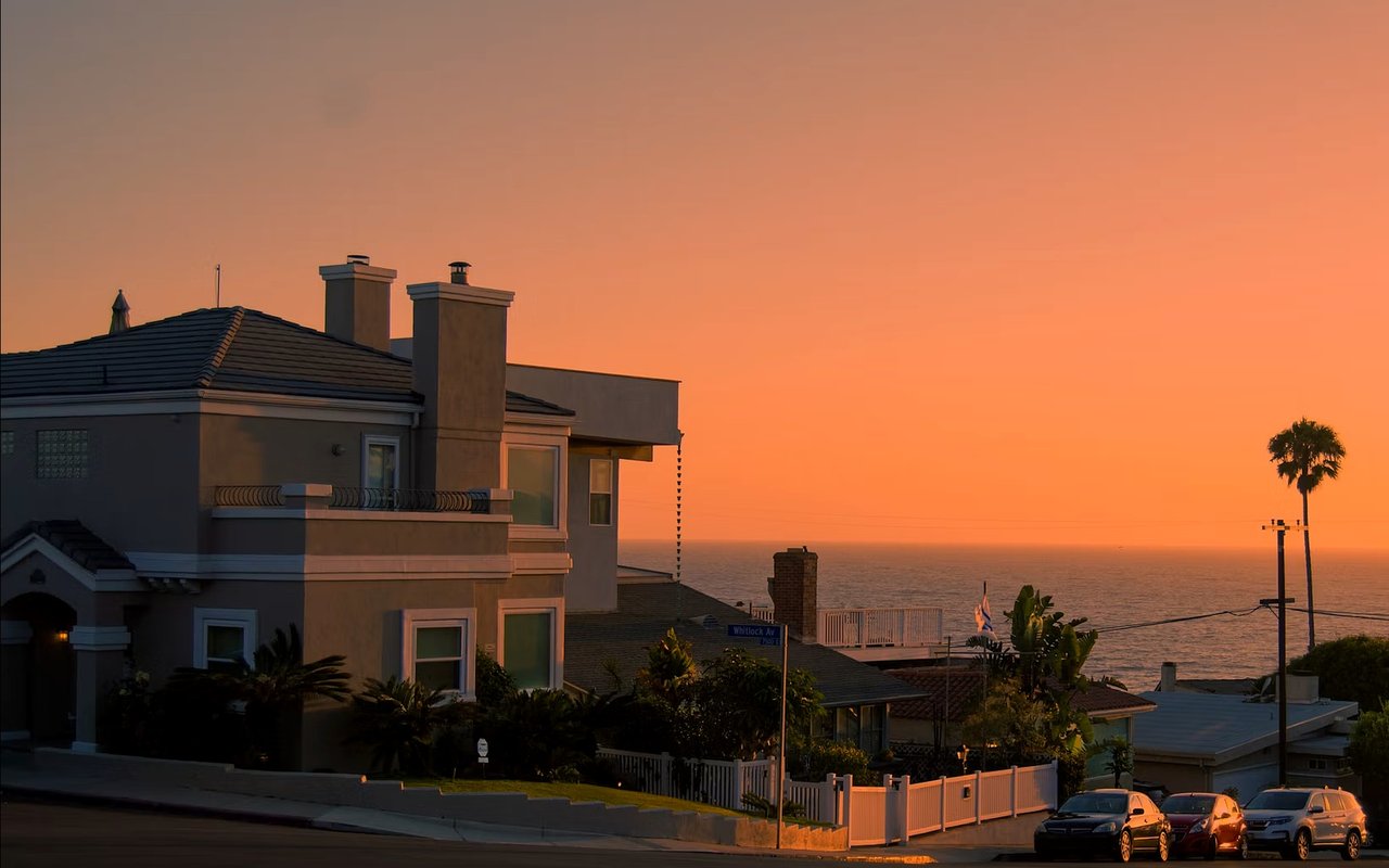 Coastal homes overlook the ocean at sunset, with a palm tree and parked cars along the street.