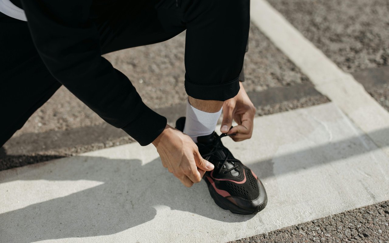 Person tying a black running shoe with red accents on a sunlit pavement.