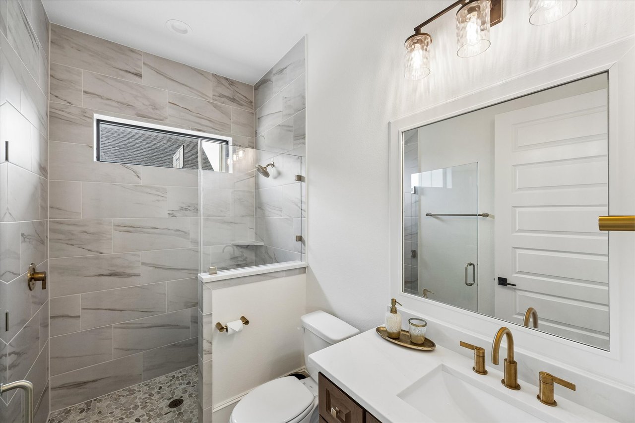Modern bathroom with a glass walk-in shower, gray tile, pebble floor, and brass vanity fixtures.