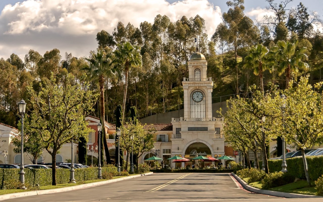 Shopping center entrance with a clock tower, palms, and outdoor dining along a tree-lined road.