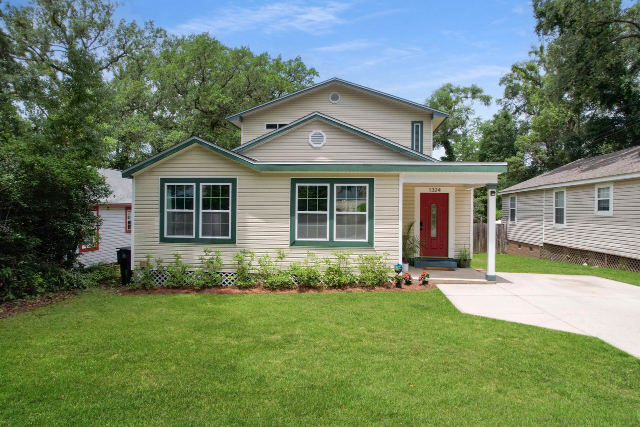 A charming, two-story white house with green trim, featuring a red door, is surrounded by lush greenery and a well-manicured lawn under a blue sky.