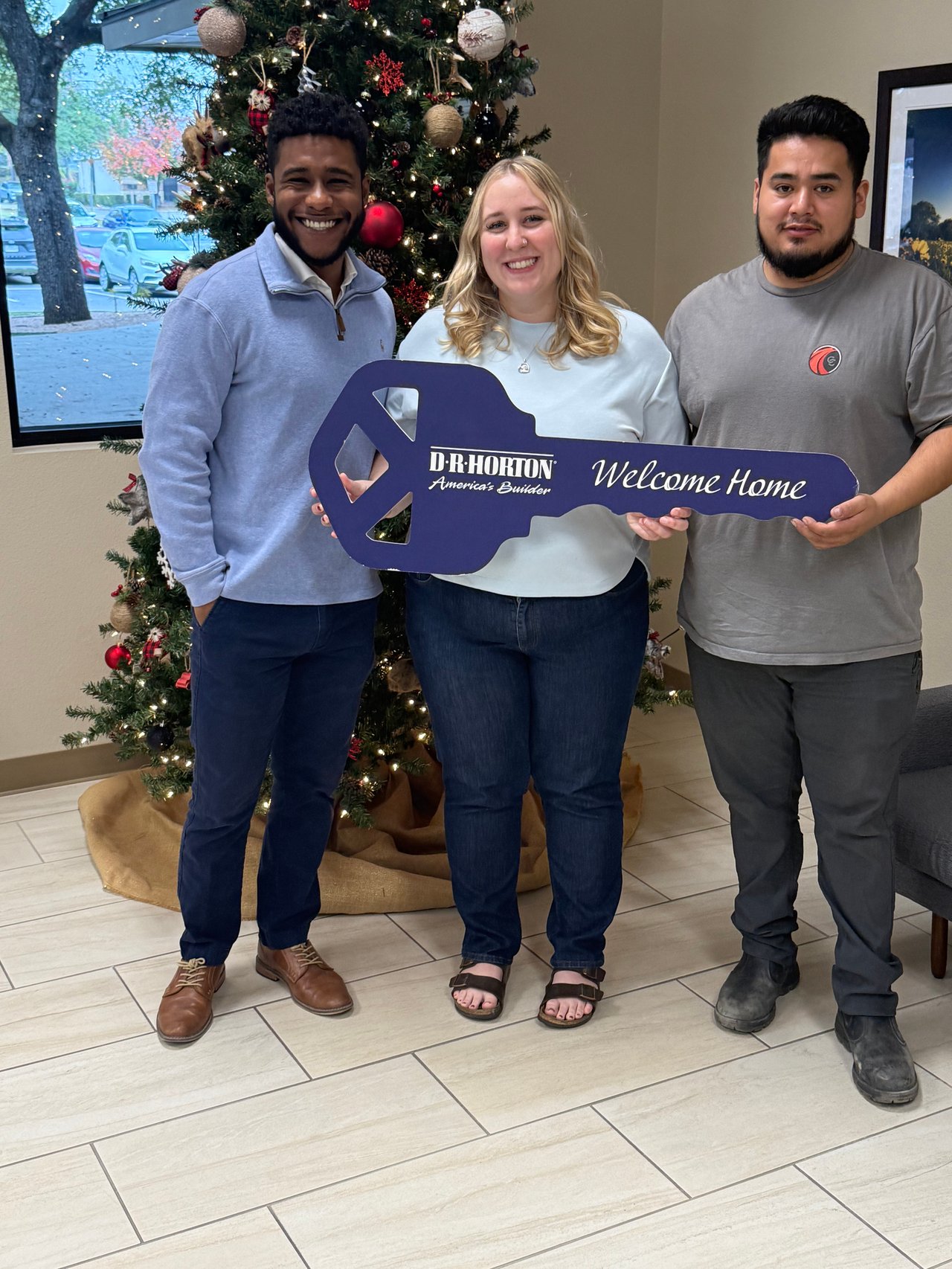 Three people pose with a D.R. Horton welcome-home key sign beside a decorated Christmas tree.