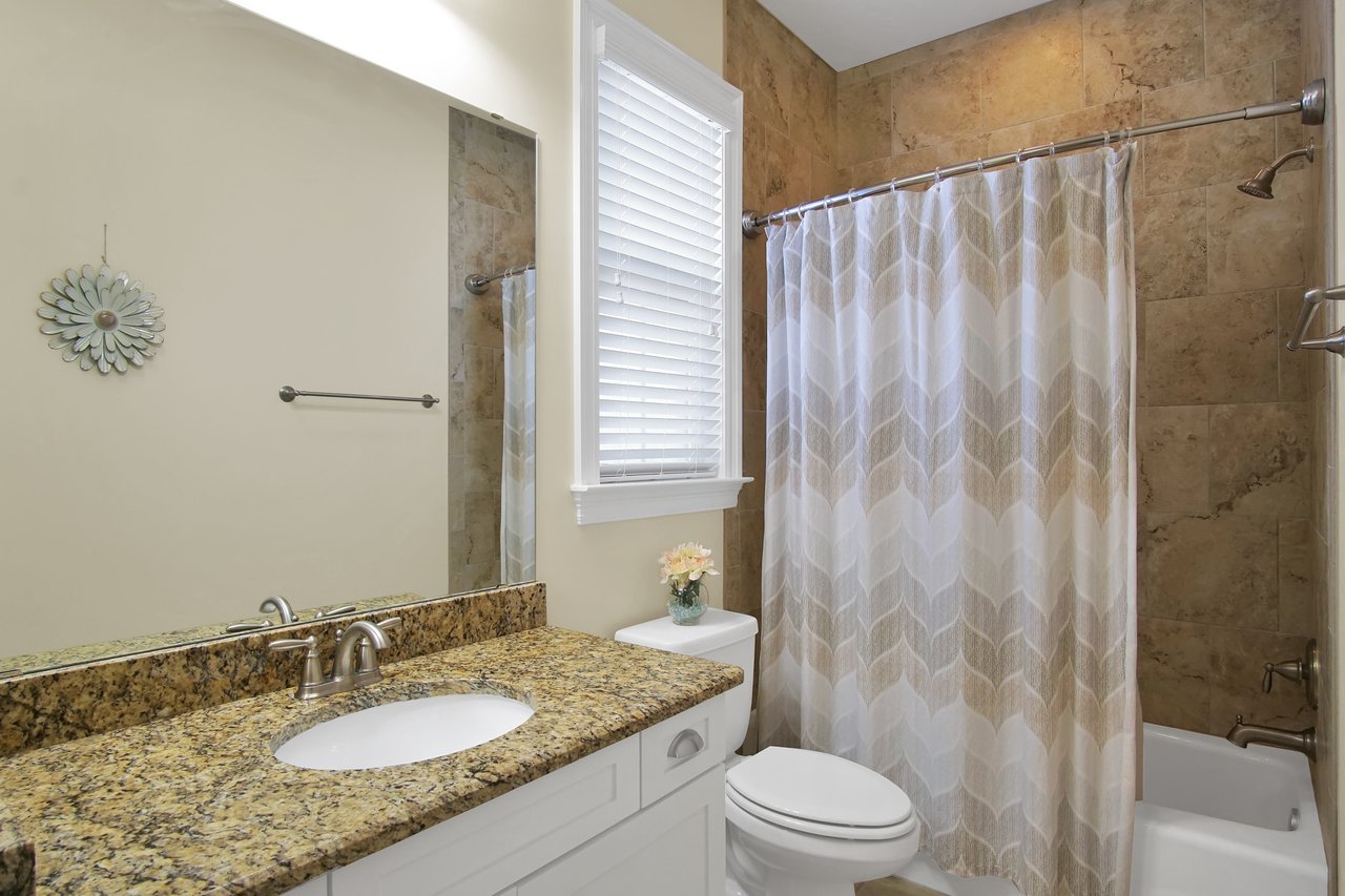 Modern bathroom with granite countertop, white cabinet, and brushed metal fixtures. Features a tiled shower, patterned curtain, and window blinds.