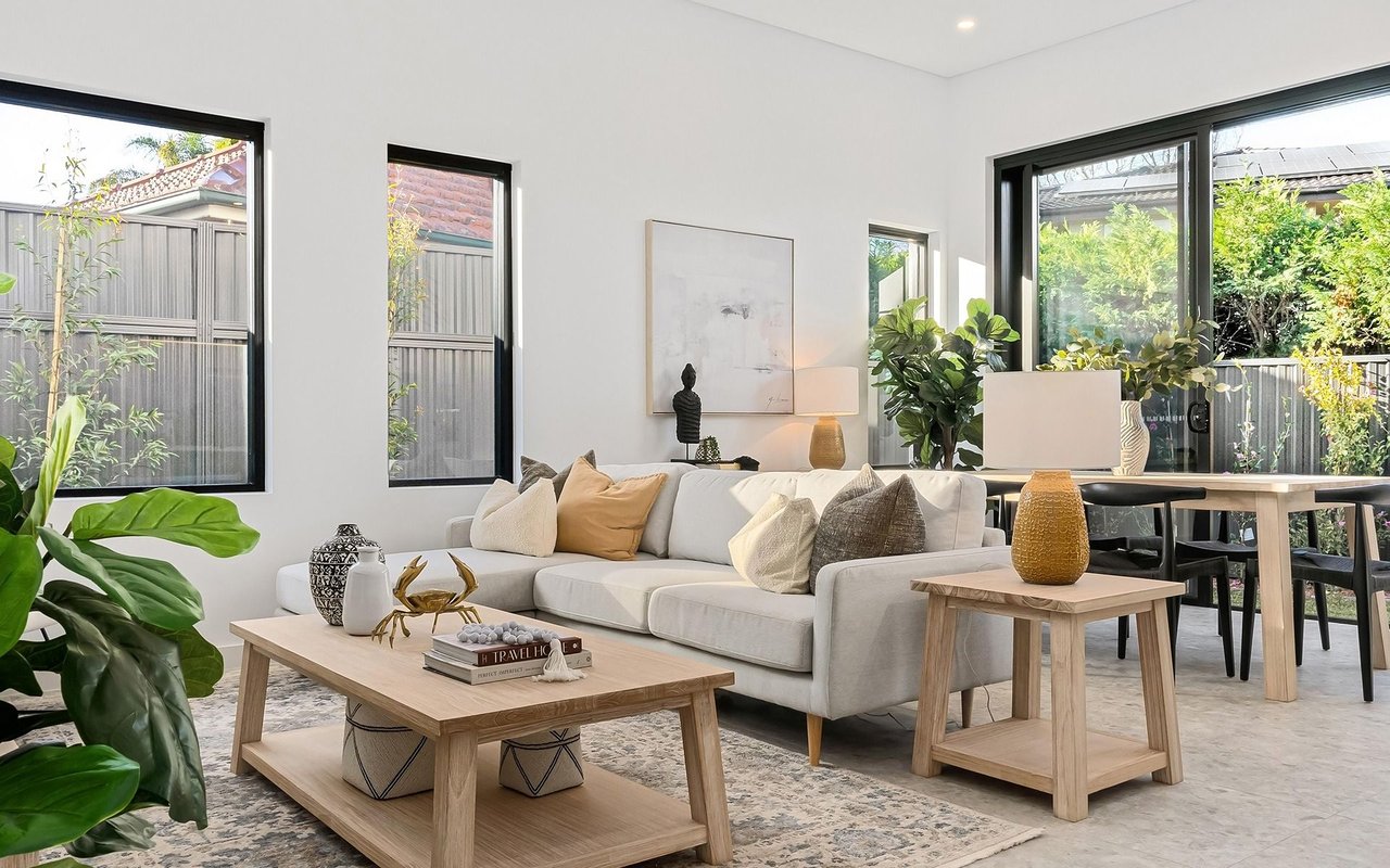 Bright living room with a white sectional, pale wood tables, and large windows overlooking greenery.