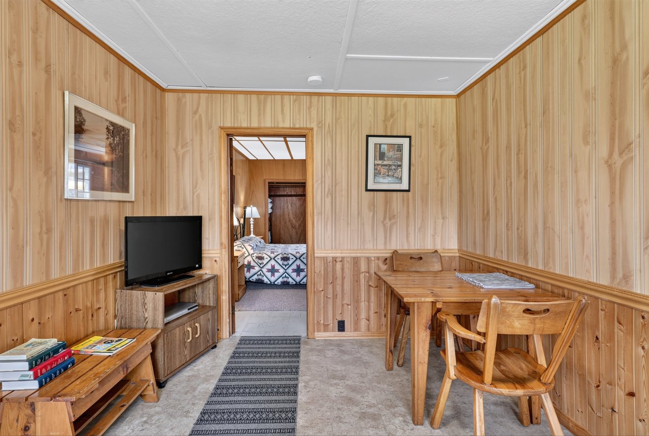 Cottage dining area with knotty-pine walls, wooden furniture, and a view into the bedroom.