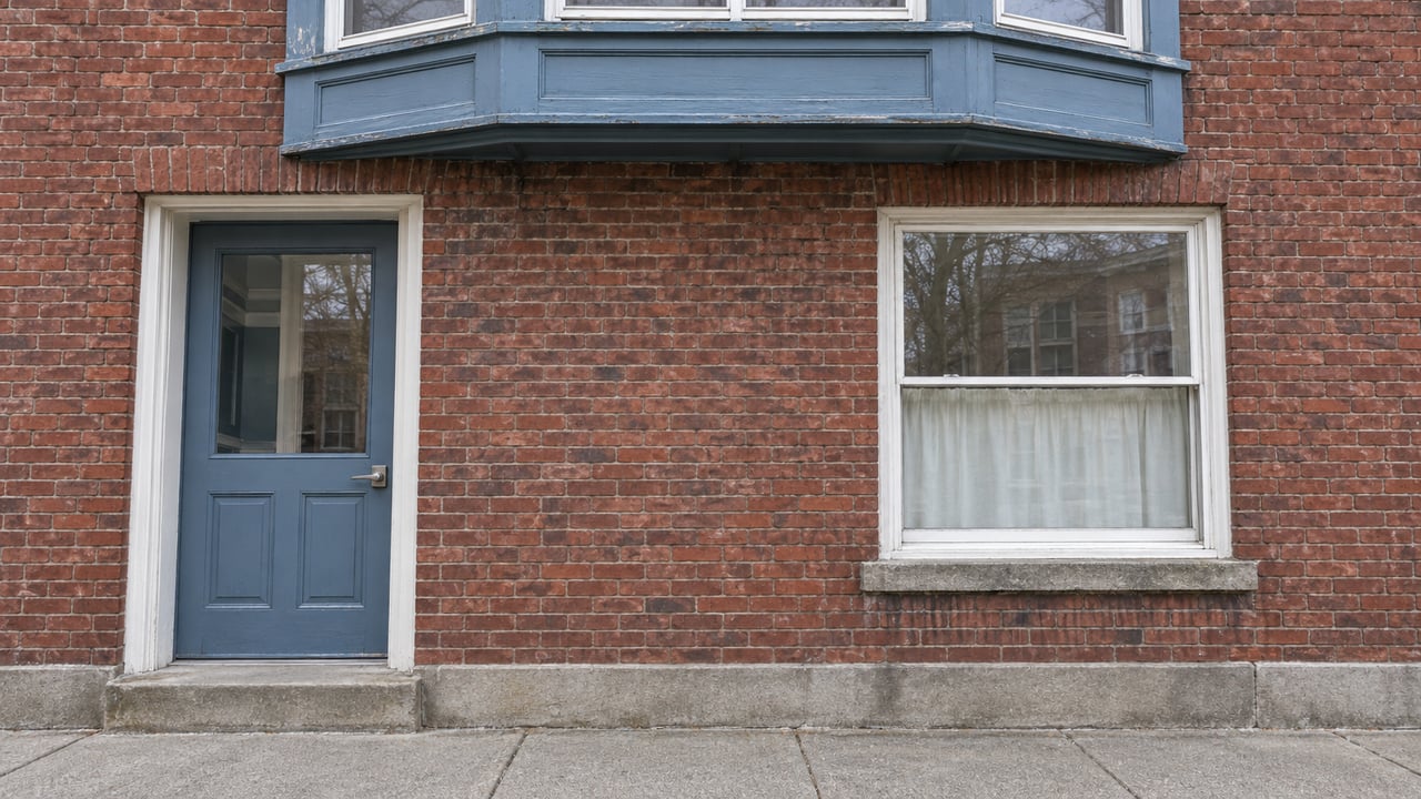 Blue-painted residential entrance and white sash windows in an aged red-brick facade above a concrete sidewalk.