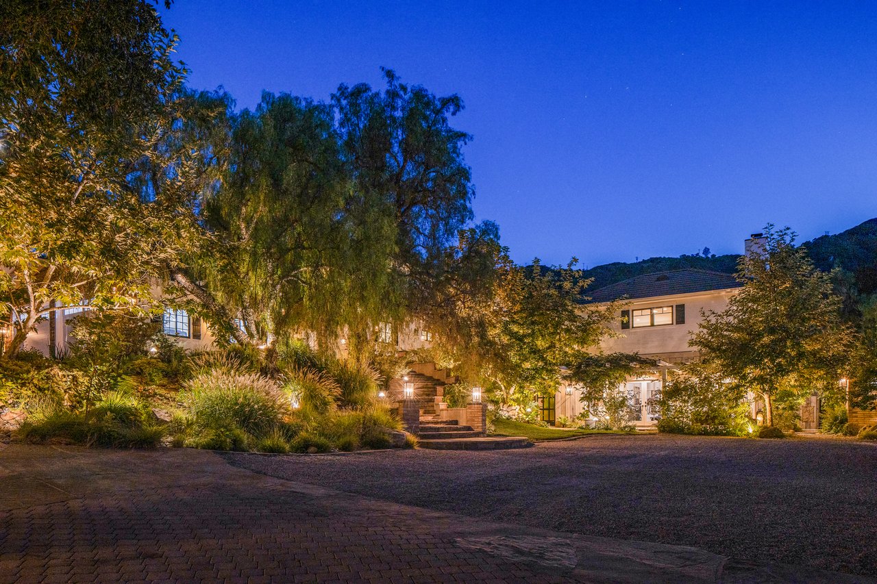 Two-story home glowing at dusk behind mature trees, with stone entry steps and a broad paved driveway.