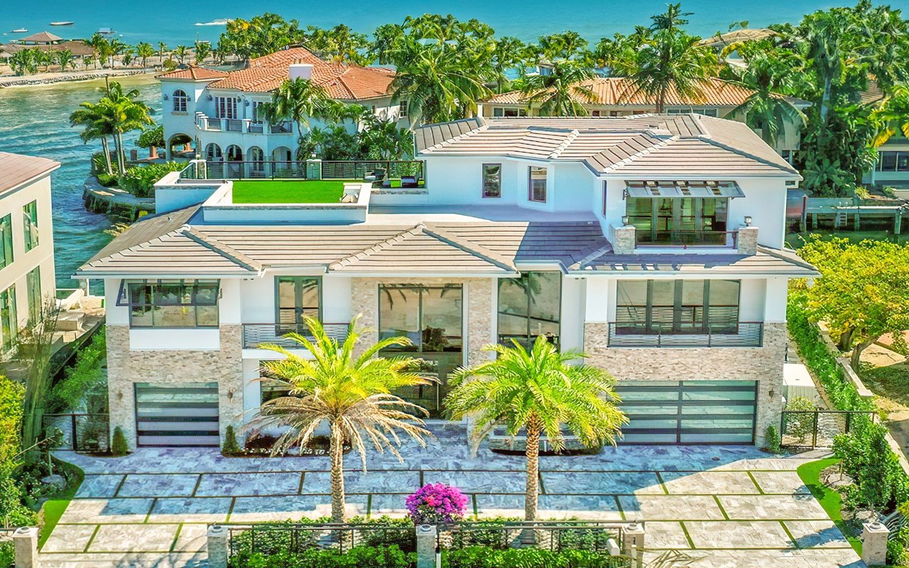 Modern waterfront home with broad tile roofs, palm-lined driveway, and neighboring homes across the waterway.
