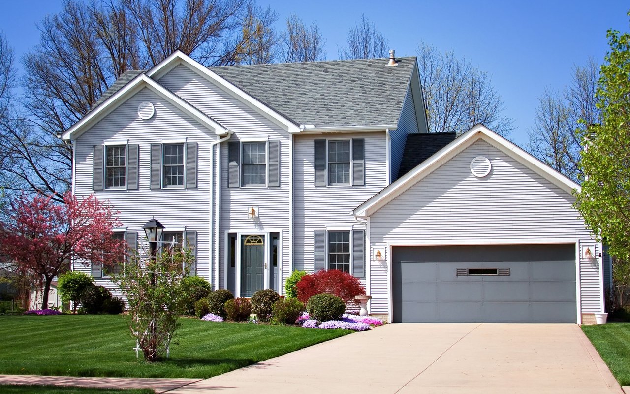 Two-story gray house with an attached double garage, landscaped lawn, and flowering tree.