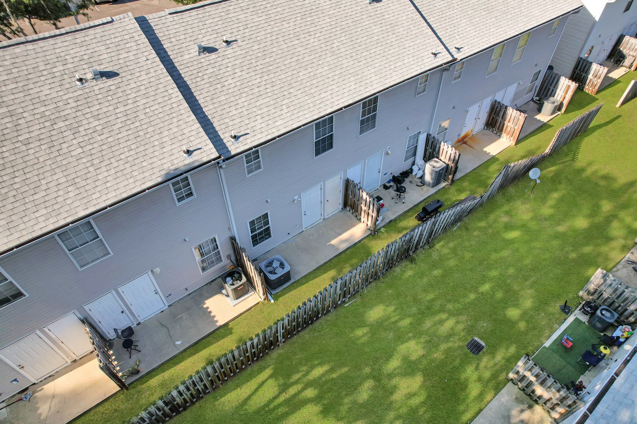 Aerial view of a row of townhouses with fenced backyards featuring paved patios and grassy areas. Objects like chairs and air conditioning units are visible. The scene conveys a peaceful residential vibe.Aerial view of a row of townhouses with fenced backyards featuring paved patios and grassy areas. Objects like chairs and air conditioning units are visible. The scene conveys a peaceful residential vibe.