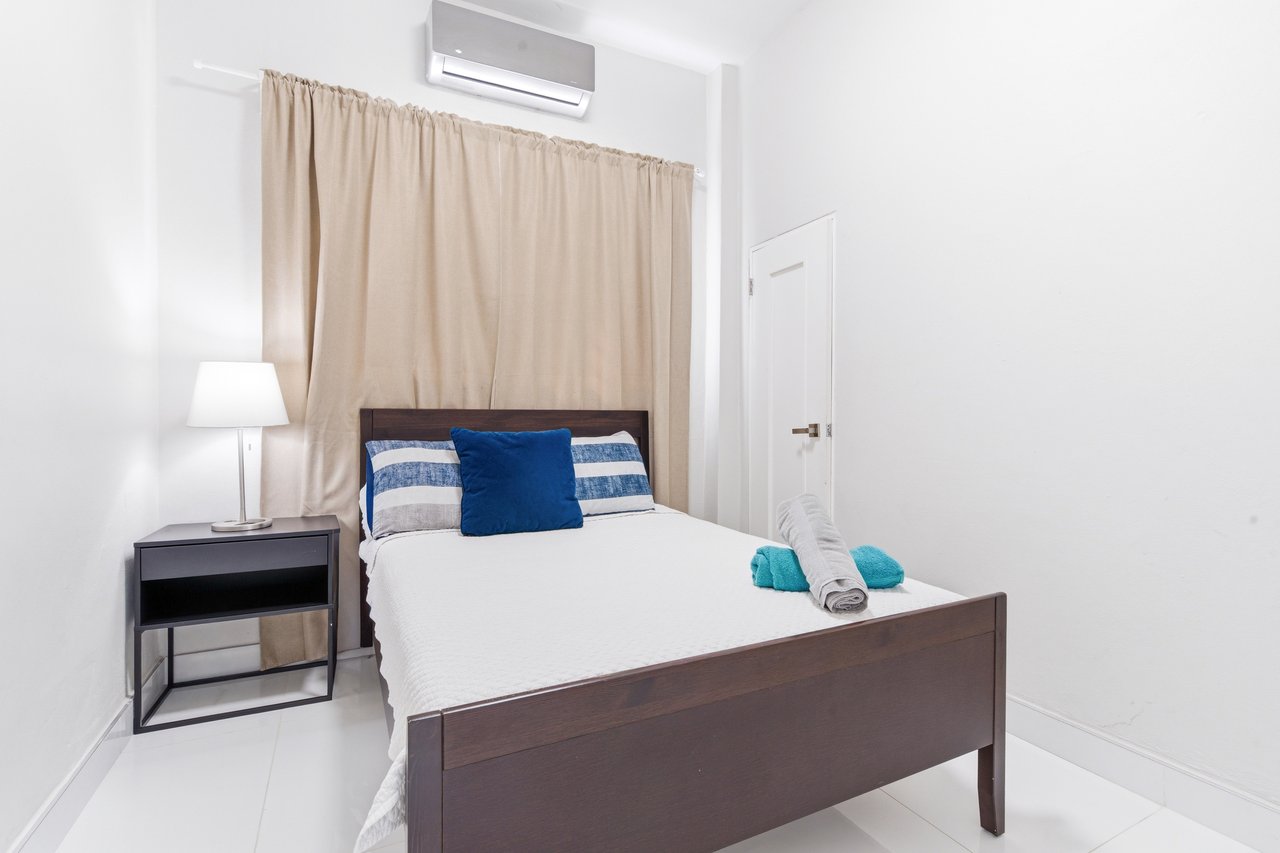 Bedroom with a dark wooden bed, blue accent pillows, beige curtains, and a wall-mounted air conditioner.
